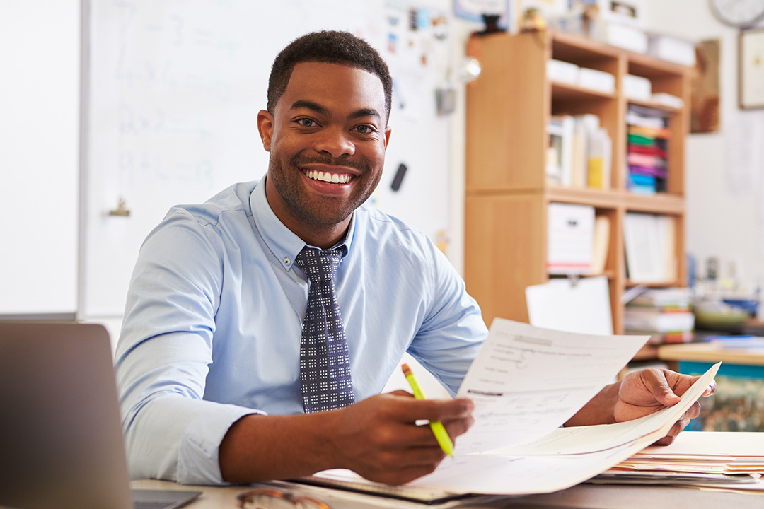 Male teacher, sitting at his desk, marking students work