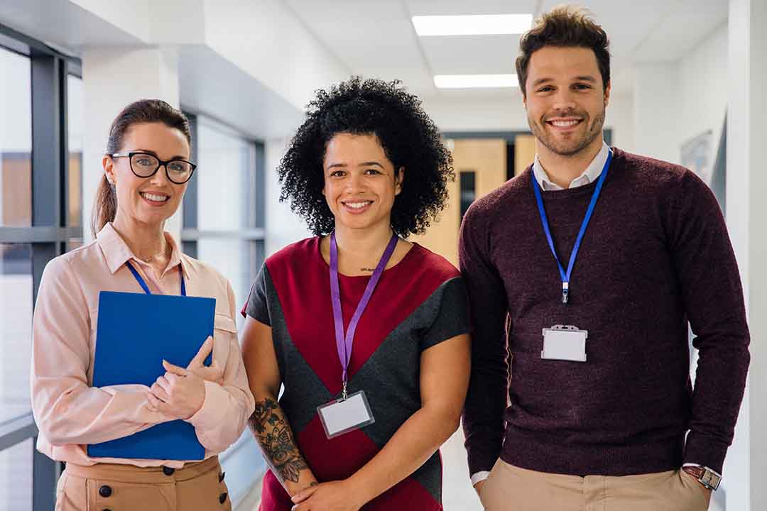 Three teachers standing side-by-side smiling at the camera, in a school corridor.