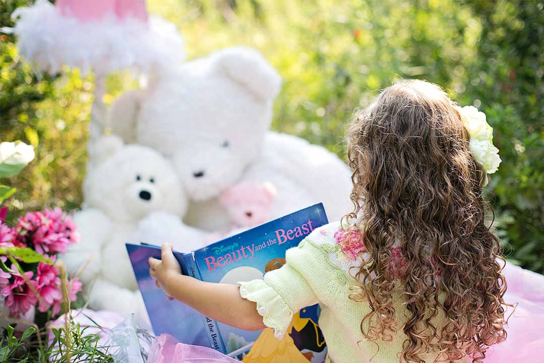 A little girl, sitting outside, on a picnic blanket, reading a book to her two fluffy teddy bears.