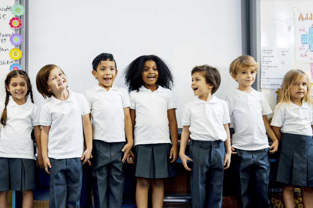 7 smiling children in white shirts standing in front of a white board